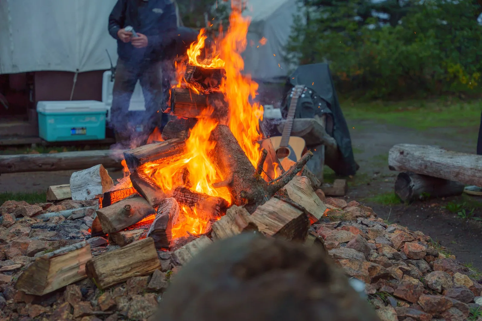 Campfire blazing at dusk in front of the wall tents with a guitar resting nearby